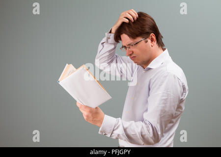 Un giovane uomo, incentrato sulla lettura di un libro nel coperchio bianco, con confusa emozione sulla faccia, graffi la sua testa; orientamento orizzontale ritratto in studio. Foto Stock