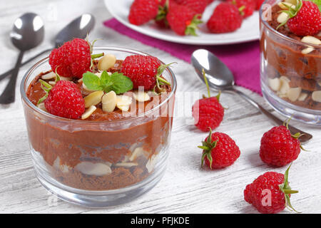 Deliziosa mousse al cioccolato in bicchieri di vetro e rabboccato con foglie di menta fresca, lamponi e scaglie di mandorle sulla tavola con cucchiai su sfondo, vista da ab Foto Stock