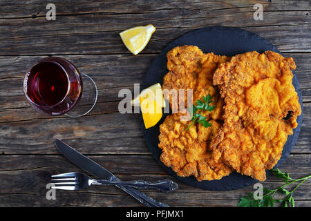 Deliziosa golden brown classic Wiener Schnitzel - o panate cotolette servita sulla pietra nera piastra con un bicchiere di vino rosso scuro sul tavolo di legno, vie Foto Stock