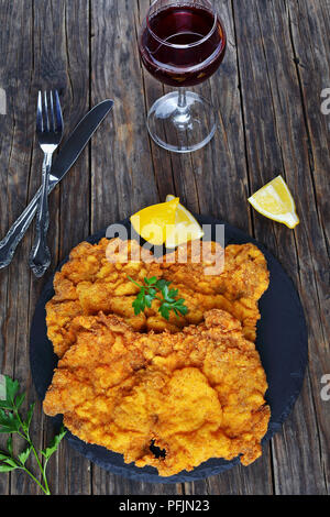 Deliziosa golden brown classic Wiener Schnitzel - o panate cotolette servita sulla pietra nera piastra con un bicchiere di vino rosso scuro sul tavolo di legno, ver Foto Stock