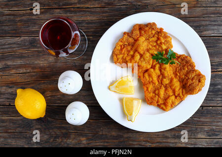 Deliziosa golden brown classic Wiener Schnitzel - o panate cotolette servita su piastra bianca con le fette di limone su dark tavolo in legno con vetro di r Foto Stock