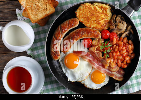 Close-up di colazione inglese completa - fagiolo, hash brown, uova fritte, pancetta, salsicce arrostite, pomodori, funghi sulla padella scuro sul tavolo di legno con t Foto Stock