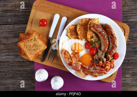 La completa prima colazione inglese - fagiolo, hash brown, uova fritte sunny side up, fettine di pancetta, salsicce, pomodori e funghi sulla piastra bianca sul bordo di taglio con Foto Stock