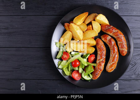 Deliziosi hot salsicce alla griglia, patate al forno e verdure fresche romaine insalata di lattuga sul piatto nero su nero di tavole di legno, vista da sopra Foto Stock