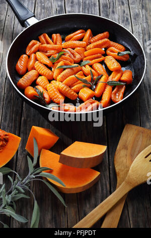 Stuzzicanti gnocchi di zucca fritto nel burro con foglie di salvia fresca in una padella. I pezzi di zucca e spatole in legno sul vecchio legno scuro, tabella vertic Foto Stock