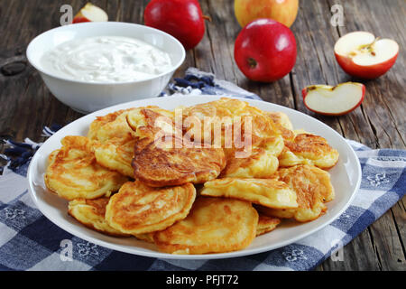 Deliziose frittelle fatte in casa caricato con succosi pezzi di mela, sulla piastra bianca sul vecchio legno scuro tavolo con panna acida in un recipiente e le mele a backgroun Foto Stock
