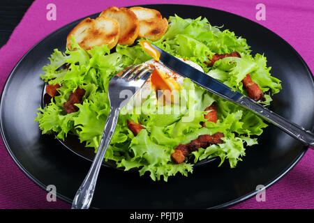 Frisee lattuga, pancetta croccante e un uovo in camicia - insalata fresca Lyonnaise deliziosa insalata, servita su piastra nera con crostini di baguette, autentica destinatario Foto Stock