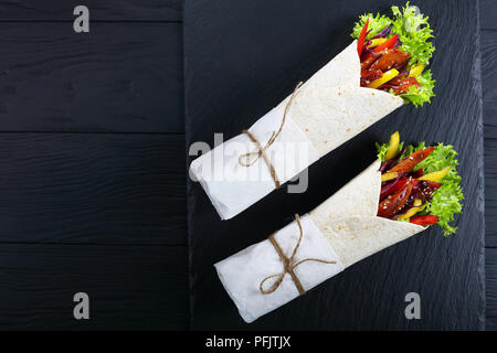 Tortilla arrotolato attorno ad un riempimento - rosso insalata di cavolo, il peperone e le strisce di pollo fritto carni in ardesia nera, vassoio sul tavolo di legno, vista da abov Foto Stock