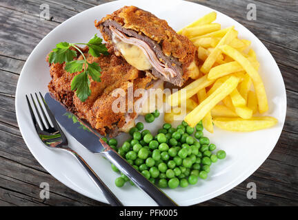 Deliziosa cotoletta Cordon Blue con patatine fritte, piselli verdi e prezzemolo sulla piastra sul vecchio legno scuro tabella d'argento con coltello e forchetta, autentica ricetta, Foto Stock