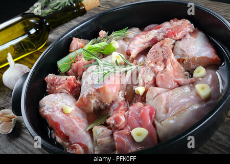 Pezzi di coniglio crudo preparare per essere cotti, marinato con vino bianco, aglio e bouquet garni in nero teglia sul tavolo con bottiglia di vino e Foto Stock