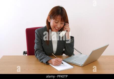 Sorridente imprenditrice sitiing alla scrivania, utilizzando il telefono e la scrittura della nota sul tastierino accanto al laptop Foto Stock