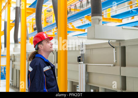 Lavoratore in una fabbrica che indossa blu di lavoro tuta e casco rosso cercando in macchine in background Foto Stock