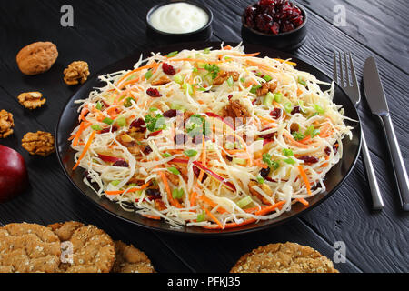 Close-up di Apple di mirtillo palustre e noci insalata Coleslaw su una piastra nera su tavoli in legno nero con ingredienti e fette biscottate a sfondo, vista dal Foto Stock