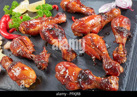 Yummy grigliate di croccante succosa pollo teriyaki bacchette cosparso con i semi di sesamo sulla pietra nera vassoio sul tavolo di legno con prezzemolo, aglio e limone Foto Stock