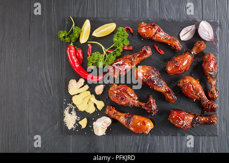 Yummy grigliate di croccante succosa pollo teriyaki bacchette cosparso con i semi di sesamo sulla pietra nera vassoio sul tavolo di legno con prezzemolo, aglio e limone Foto Stock