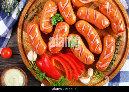 Alla griglia calda succosa salsicce di manzo con timo fresco sul round tagliere in legno scuro tavolo da cucina con peperone rosso, ketchup e basic aioli sau Foto Stock