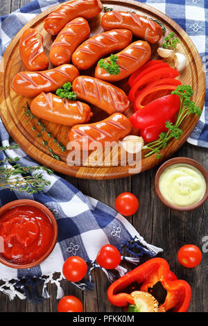 Alla griglia calda succosa salsicce di manzo con timo fresco sul round tagliere in legno scuro tavolo da cucina con peperone rosso, ketchup e basic aioli sau Foto Stock