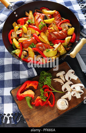 Hot deliziose salsicce alla griglia con verdure, funghi e prezzemolo in una padella, ingredienti sul bordo di taglio verticale, vista da sopra Foto Stock