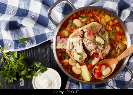 Deliziosa carne di maiale minestra di fagioli bianchi, i piselli, il peperone rosso, erbe, specie in una casseruola di metallo con il cucchiaio di legno, su di un legno nero Foto Stock