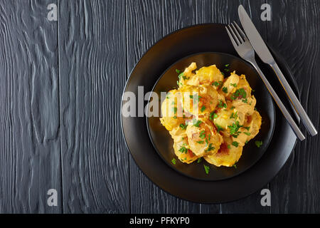 Cheesy patate novelle con il prezzemolo tritato servite su piastre nere con argento forchetta e coltello su un tavoli in legno nero, vista da sopra, vicino-u Foto Stock