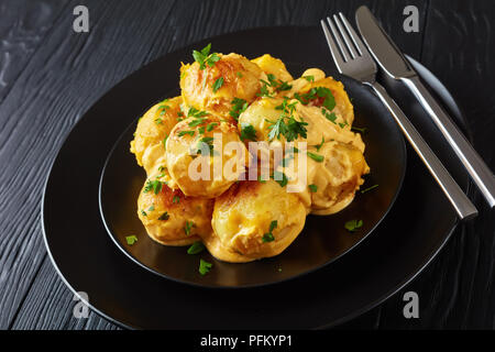 Cheesy patate novelle con il prezzemolo tritato servite su piastre nere con argento forchetta e coltello su un tavoli in legno nero, vista da sopra, vicino-u Foto Stock