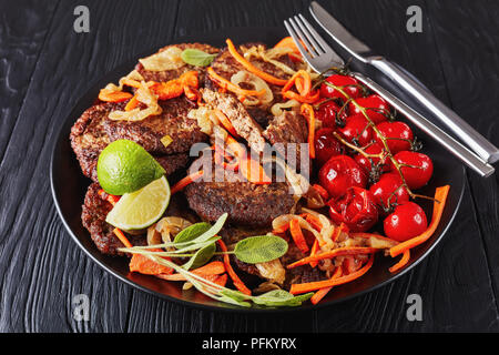 Fegato di pollo frittelle con carota, cipolla, pomodoro grigliate servita su una piastra nera con coltello e forchetta, orizzontale vista da sopra Foto Stock