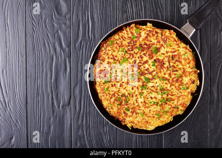 Swiss röstinchen o potato pancake cosparso con del prezzemolo tritato finemente in una padella su tavoli in legno nero, vista orizzontale dal di sopra, laici piatta Foto Stock