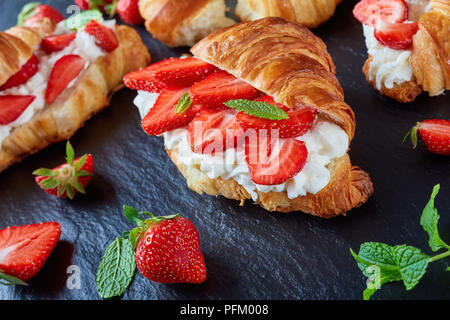 Deliziosi croissant francesi sandwich multistrato con fresche fragole mature e panna montata il formaggio su un vassoio di pietra su tavoli in legno nero, vista dal Foto Stock