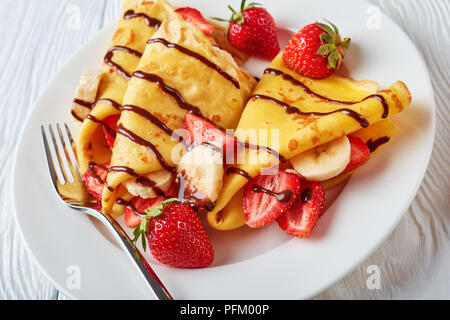 Frittelle sottili o crepes su una piastra bianca con fragole e fette di banana e crema di cioccolato, vista orizzontale dal di sopra, close-up Foto Stock