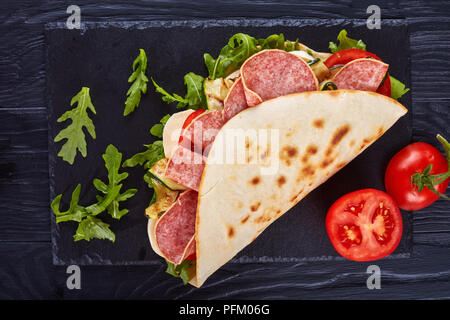 Un ragazzo caucasico con una camicia taglia la pizza con il salame con un  coltello tondo in cucina sul tavolo pizza tonda con pomodori e salame su  uno sfondo di legnofast food