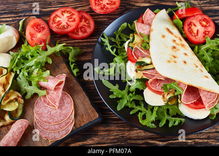 Aprire il panino - italiano piadina con mozzarella, pomodoro, fette di salame, zucchine grigliate e rucola su una piastra nera con ingredienti su una scheda in legno Foto Stock