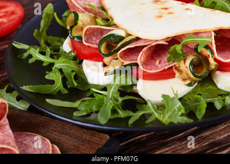 Aprire il panino - italiano piadina con mozzarella, pomodoro, fette di salame, zucchine grigliate e rucola su una piastra nera con ingredienti su un taglio di bo Foto Stock