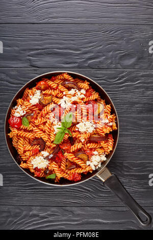 La pasta italiana I fusilli alla Norma con melanzane, pomodori e basilico, ricotta in una padella su tavoli in legno nero, verticale vista da sopra, vicino- Foto Stock