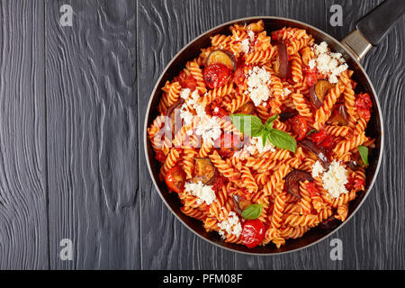 La pasta italiana I fusilli alla Norma con melanzane, pomodori e basilico, ricotta in una padella su tavoli in legno nero, vista orizzontale dal di sopra, clos Foto Stock