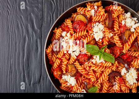 Close-up di pasta italiana fusilli alla Norma con melanzane, pomodori e basilico, ricotta in una padella su tavoli in legno nero, vista orizzontale da Foto Stock