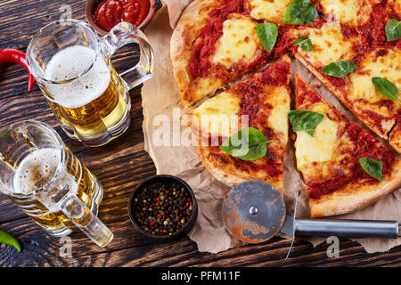 Close-up di italiano classico Pizza Margherita tagliato a fettine su una carta pergamena su un vecchio rustico tavolo in legno con la birra in bicchieri di vetro, autentico reci Foto Stock