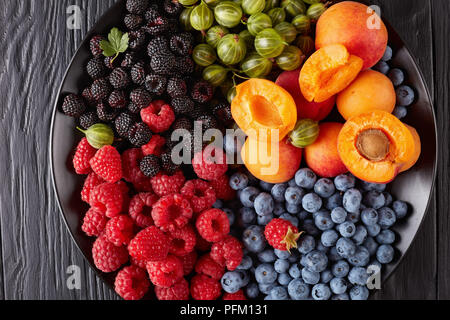 Close-up di frutti e bacche insalata di organico fresco Ribes rosso e nero lampone, mirtillo, fettine di albicocche su una piastra nera su una tavola di legno Foto Stock
