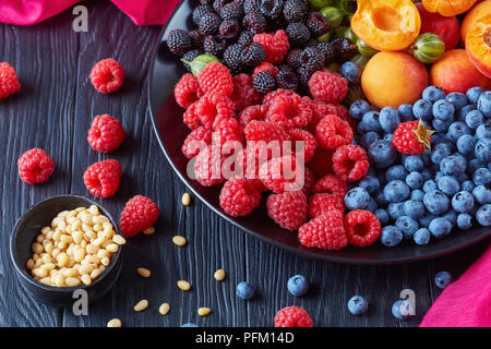 Close-up di frutti e bacche insalata con organici freschi ribes rosso e nero lampone, mirtillo, fettine di albicocche su una piastra nera su una scheda in legno Foto Stock