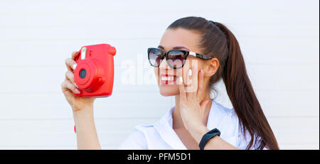 Moda ritratto di bella donna sorridente in occhiali da sole rendendo la foto dalla fotocamera contro il muro grigio. Cuffie wireless, fitness bracciale sul lato Foto Stock