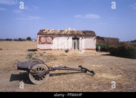 India, Sawai Madhopur, carrello di legno in piedi sul secco di massa terrosa, villaggio bianco Rifugio con la paglia su semplice tetto di tegole, dipinto sulla parete di capanna, Capra in piedi accanto a capanna, campo in background. Foto Stock
