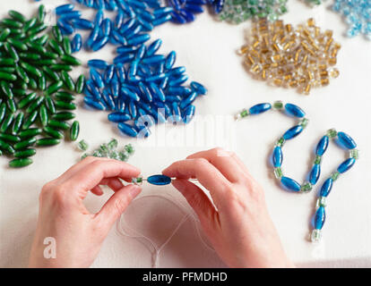 Mani blu di threading tallone sopra l'ago sul filo bianco, filettato il blu e il verde delle perline di vetro la raccolta a fine, pile di blu, verde e ambra di perline in background. Foto Stock