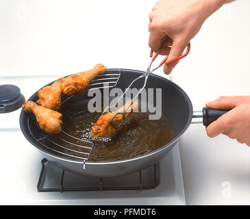 Mani servendosi di un paio di pinze per spostare fritte coscia di pollo dal tegame su scolapiatti Foto Stock