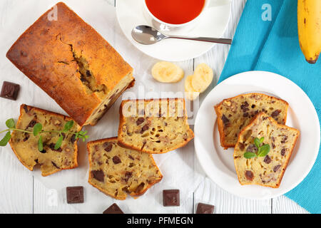 Close-up di pane appena sfornato delizioso pane alla banana con noci e cioccolato di pezzi tagliati in fettine su carta. ingredienti e la tazza di tè sul tavolo di legno, Foto Stock