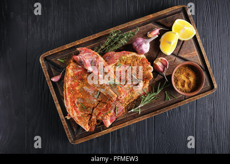 Crudo fresco di ricambio costolette di agnello marinato e spolverati con un mix di spezie di terra su legno marrone tagliere con rosmarino, timo, aglio e fetta di limone Foto Stock