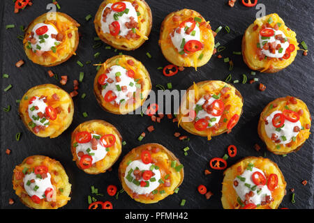 Patate cotte due volte metà caricato con formaggio Cheddar grattugiato, pancetta, peperoncino fette, panna acida e cosparsi di cipolla verde sul vassoio di ardesia, o Foto Stock