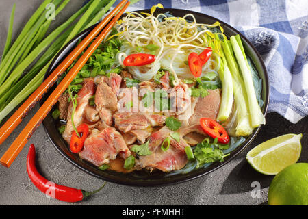 Zuppa vietnamita Pho Bo con riso noodle, materie Lombata di manzo a fette, germogli di soia, calce, cipolline in nero ciotola con bacchette e ingredienti sul co Foto Stock