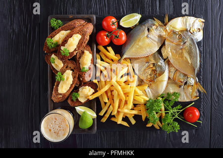 Deliziosa freddo affumicato luna Pesce e Patatine serviti su un nero ardesia vassoio con il capelin caviale toast, calce e bicchiere di birra, vista da sopra, close-up Foto Stock