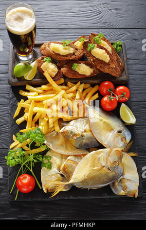 Deliziosa freddo affumicato pesce e patatine fritte servite su un nero ardesia vassoio con il capelin caviale pane tostato e un bicchiere di birra, verticale vista da sopra Foto Stock