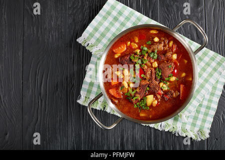 Delizioso caldo gulasch ungherese con carne di manzo, paprica, verdure e csipetke - piccolo uovo tagliatelle in acciaio inox pentola su tavoli in legno nero, il CLA Foto Stock