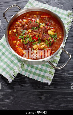 Delizioso caldo gulasch ungherese con carne di manzo, paprica, verdure, csipetke, spezie in acciaio inox pentola su tavoli in legno nero, autentica ricetta, Foto Stock